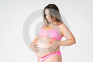 Happy plus size pregnant woman in pink underwear applying skin stretch mark cream on her belly on gray background, maternity