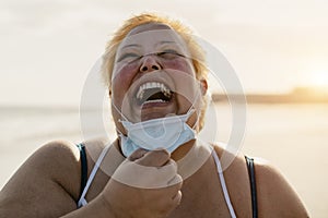 Happy plus size woman laughing on the beach while wearing face mask - Curvy overweight model having fun during vacation