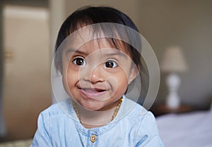 Happy, portrait smile and Down syndrome baby relaxing on a bed in happiness at home. Cheerful little child with genetic