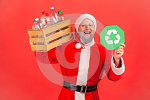 Happy positive elderly man with gray beard wearing santa claus costume holding box with plastic bottles on shoulder and showing