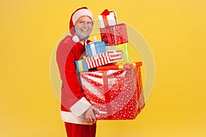 Happy positive elderly man with gray beard wearing santa claus costume posing with stack of presents