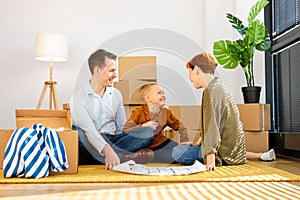 happy positive family with child boy sitting on floor among cardboard boxes in new house, using blueprint