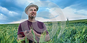Happy positive farmer in hat standing in wheat field and calculating on tablet