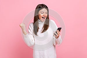 Happy positive girl holds smart phone in hands, clenching fist, celebrating victory, sudden success.