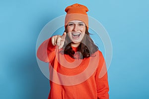 Happy positive laughing female wearing jumper and hat pointing finger to camera and makes fun of somebody, being in good mood,