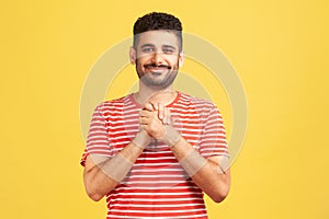 Happy positive man with beard in striped shirt pressing hands together on chest enjoying, calming and comforting himself