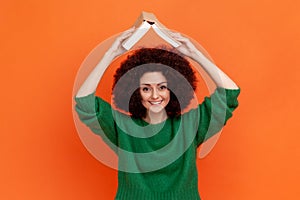 Happy positive woman with Afro hairstyle wearing green casual style sweater making roof, holding