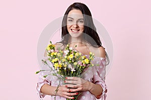 Happy positive woman with dark hair, holds flowers in hands, smiles positively, enjoys spring warm day, dressed in stylish polka