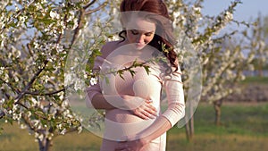 Happy pregnancy, young pregnant woman stroking belly with future baby during walk in blooming garden