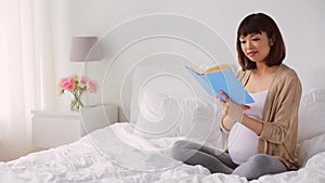 Happy pregnant asian woman reading book in bed