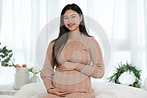Happy Pregnant Asian Woman sitting on a bed holding and stroking her big belly at home. Pregnancy of young woman enjoying with