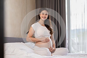 Happy Pregnant Asian Woman sitting on bed holding and stroking her big belly at home,Pregnancy of young woman enjoying with future