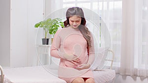 Happy Pregnant Asian Woman sitting on bed holding and stroking her big belly at home.Pregnancy of young woman enjoying with future