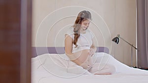 Happy Pregnant Asian Woman sitting on bed holding and stroking her big belly at home,Pregnancy of young woman enjoying with future