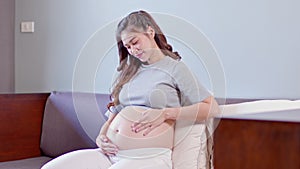 Happy Pregnant Asian Woman sitting on bed holding and stroking her big belly at home,Pregnancy of young woman enjoying with future