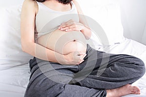 Happy pregnant asian woman sitting on bed and touching her big belly