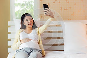 Happy pregnant asian woman taking selfie on smartphone in bed at home or Young asia pregnancy mother showing big abdomen to her
