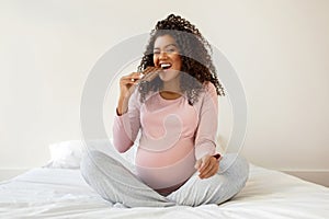 Happy pregnant black woman enjoying chocolate while sitting on bed at home