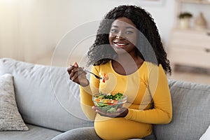 Happy pregnant black woman sitting on couch, eating fresh salad