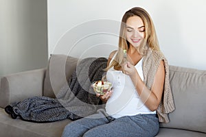 Happy pregnant blong young woman sitting on sofa eating bowl of fresh fruit  grey color of sofa and white top