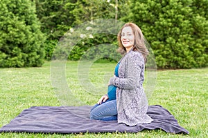 Happy pregnant caucasian woman relaxing in a park holding hands on her belly.