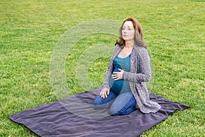 Happy pregnant caucasian woman relaxing in a park holding hands on her belly.