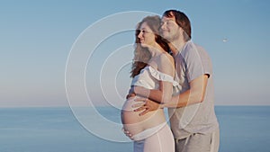 Happy pregnant couple. Husband embracing wife gently holding her belly outdoor against the sea.