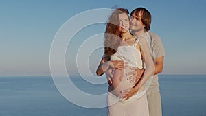 Happy pregnant couple. Husband embracing wife gently holding her belly outdoor against the sea.
