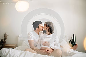 Happy pregnant couple relaxing on white bed and holding belly bump. Happy young husband kissing his smiling wife and hugging baby