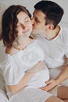 Happy pregnant couple relaxing on white bed and holding belly bump. Happy young husband kissing his smiling wife and hugging baby