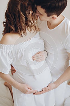 Happy pregnant couple relaxing on white bed and holding belly bump. Happy young husband kissing his smiling wife and hugging baby