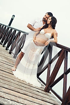 Happy pregnant couple in white clothes on the sea coast on wooden bridge