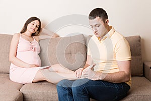 Happy pregnant family. Husband massaging his pregnant wife legs and sitting together on couch in their home