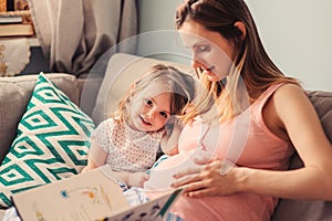 Happy pregnant mother reading book to her baby daughter at home