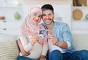 Happy pregnant muslim couple demonstrating baby sonography photo and smiling to camera