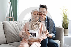 Happy Pregnant Muslim Family Sitting On Couch And Holding Baby Sonogram Picture