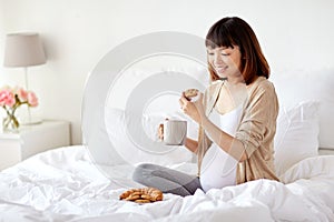 Happy pregnant woman eating cookie in bed at home