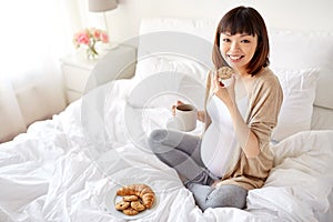 Happy pregnant woman eating cookie in bed at home