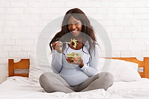 Happy pregnant woman eating fresh salad on bed at home