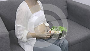 Happy pregnant woman eating fresh vegetable salad on sofa at home. Diet,