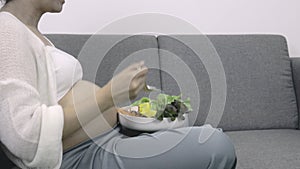 Happy pregnant woman eating fresh vegetable salad on sofa at home. Diet,