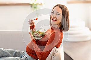 Happy Pregnant Woman Eating Salad Sitting On Couch At Home