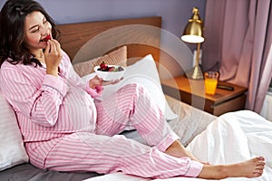 Happy pregnant woman eating strawberries on bed