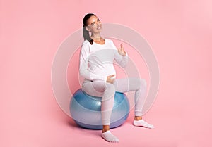 Happy Pregnant Woman Gesturing Thumbs-Up Sitting On Fitball, Pink Background