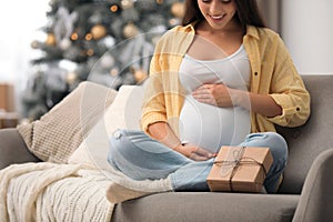 Happy pregnant woman with gift box in room decorated for Christmas, closeup. Expecting baby