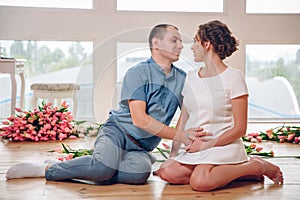 Happy pregnant woman with her husband are holding and listening to baby in belly and setting on living room`s floor at