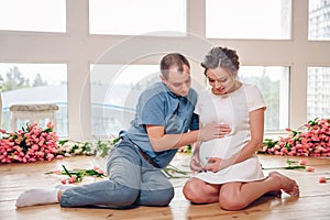 Happy pregnant woman with her husband are holding and listening to baby in belly and setting on living room`s floor at home, preg