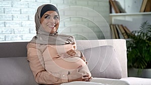 Happy pregnant woman in hijab touching belly gently, resting home, prenatal care