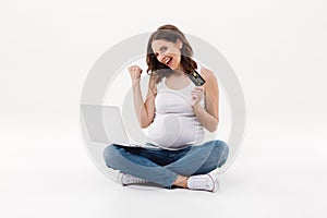 Happy pregnant woman holding debit card winking using laptop