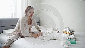 Happy pregnant woman looking at baby clothes with tenderness while sitting on sofa in domestic room. Caring future